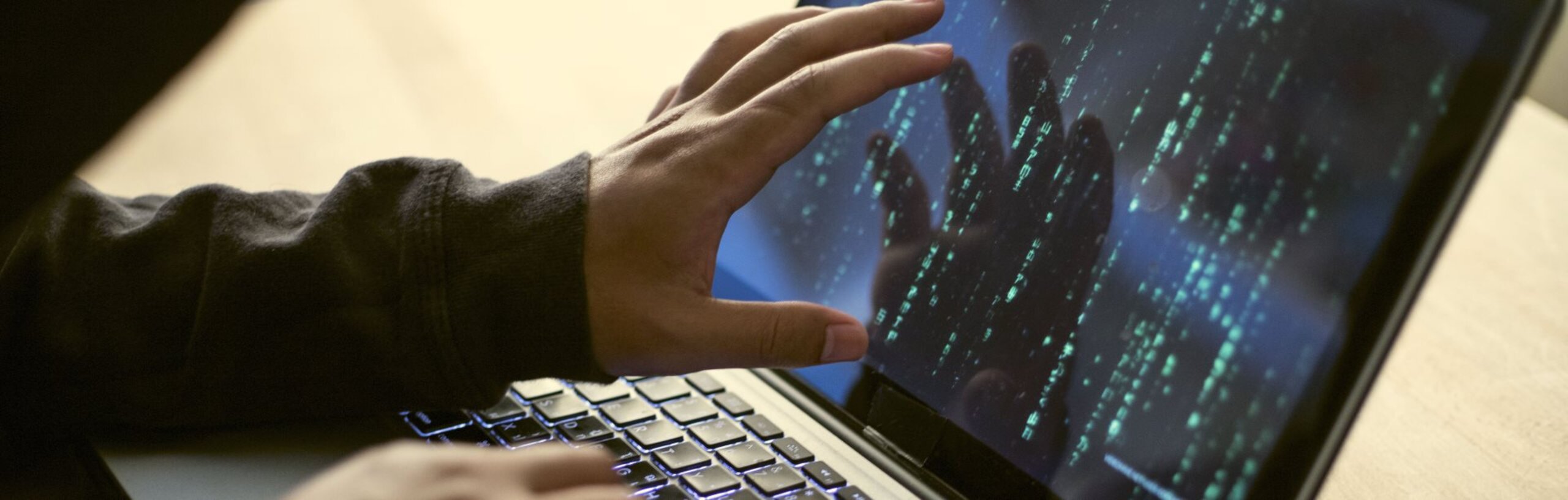 Ein Hacker sitzt an einem Laptop und zeigt mit seiner Hand auf den Bildschirm. Auf dem Bildschirm sind Zahlen und Buchstaben zu sehen.