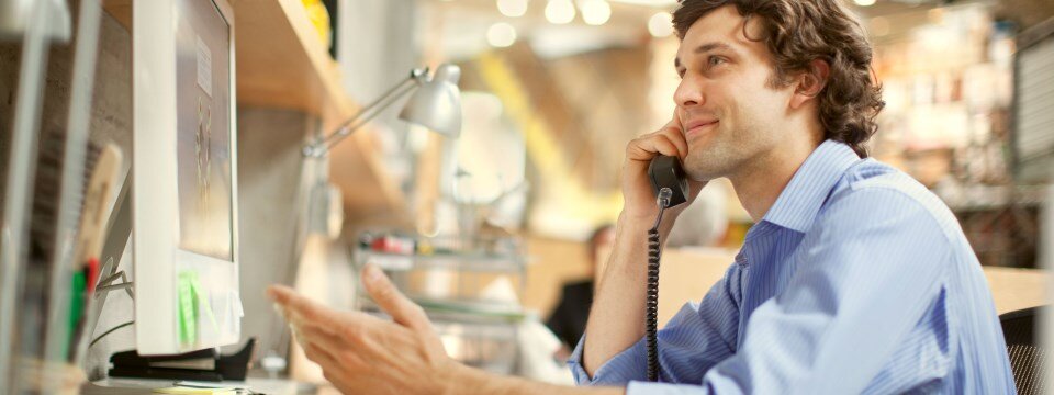 Ein junger Mann sitzt an seinem Arbeitsplatz und telefoniert mit seinem Smartphone. 