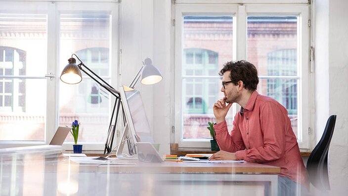 Ein Mann mit dunklen Haaren und orangenem Hemd sitzt am Schreibtisch in einem hellen Büro und guckt auf seinen Computer-Bildschirm.