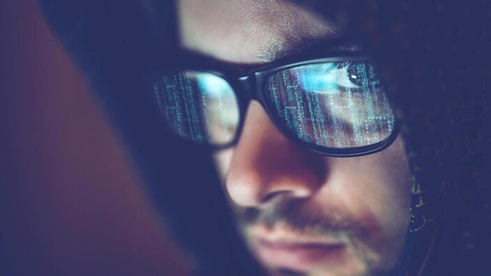 Close-up von einem Mann mit dunklem Hoodie und Brille, in der sich Computercode spiegelt.