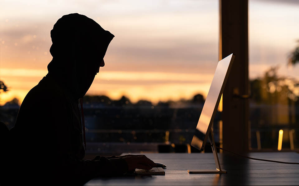 Ein Mann mit schwarzem Hoodie im Gegenlicht vor einem Computer