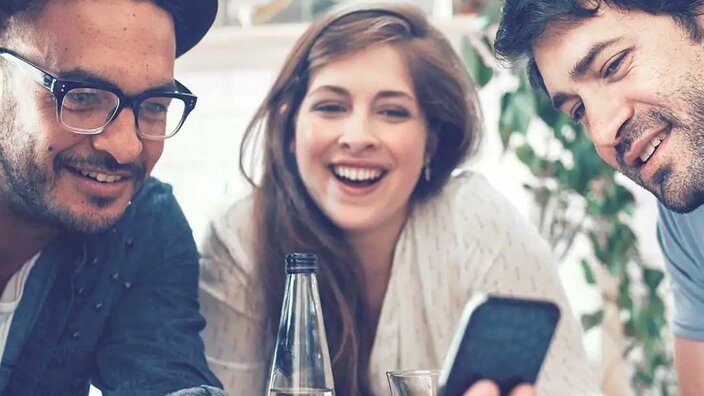 Vier junge Menschen sitzen um einen Tisch, gucken auf ein Smartphone und freuen sich.