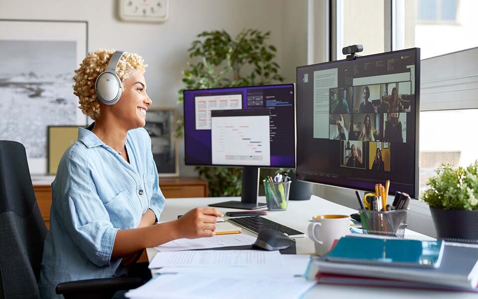 Ein blonde Frau sitzt am Schreibtisch, lächelt und nimmt an einem Online-Meeting teil, deren Teilnehmer auf ihrem Bildschirm zu sehen sind.