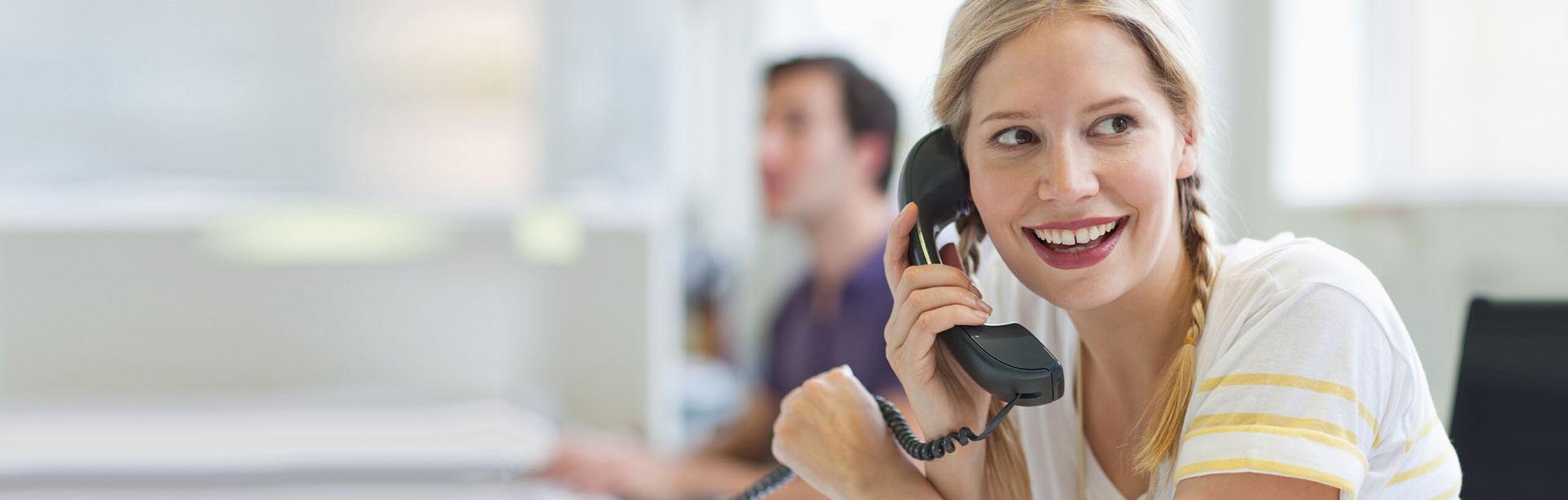Eine Mitarbeiterin im gelb-weißen Hemd telefoniert mit einem Festnetz-Telefon und lächelt.