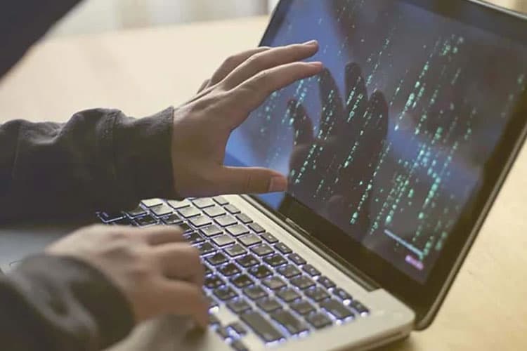 Eine Person im dunklen Hoodie bedient einen Laptop, auf dem binäre Zahlenreihen zu sehen sind.
