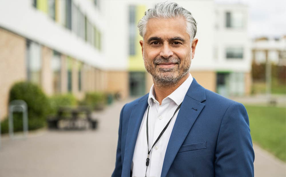 Ein Mann im blauen Jacket steht vor einem größeren Gebäudekomplex.