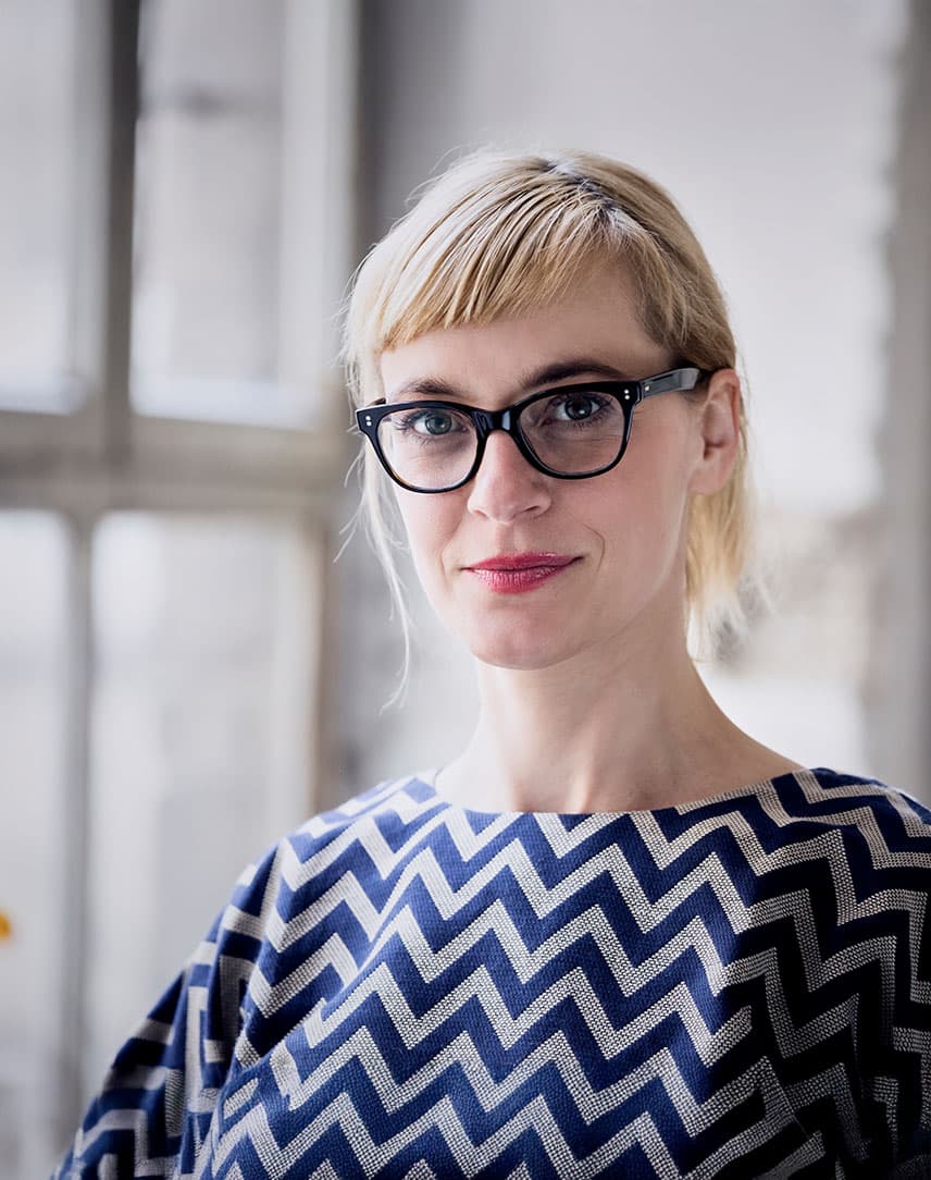 Eine Frau mit Brille guckt in die Kamera, dahinter eine Fensterfront