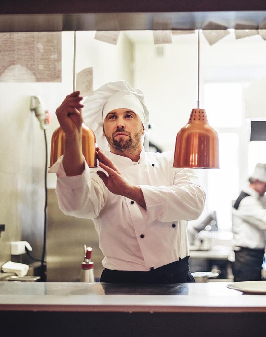 Ein Koch mit weißer Kochmütze und Schürze, der an einer Durchreiche einen Bestellzettel prüft