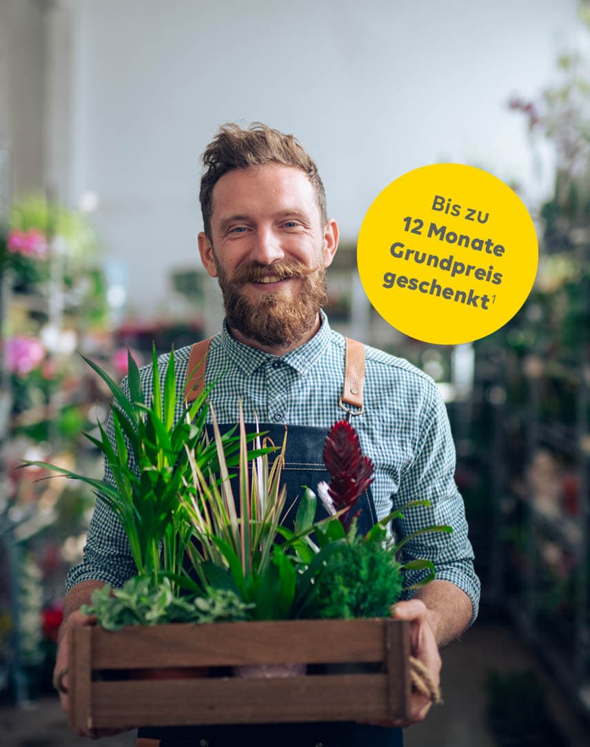 Ein Mann mit Schürze steht in einem Blumen- und Pflanzengeschäft und trägt eine Kiste mit Pflanzen in beiden Händen.