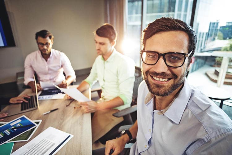 Ein Mann mit Brille an einem Bürotisch, im Hintergrund zwei Kollegen