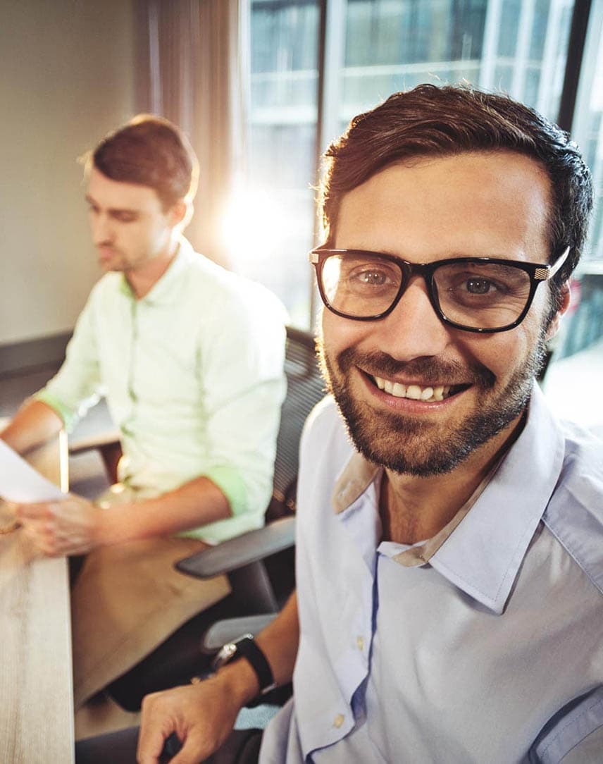 Ein Mann mit Brille an einem Bürotisch, im Hintergrund zwei Kollegen