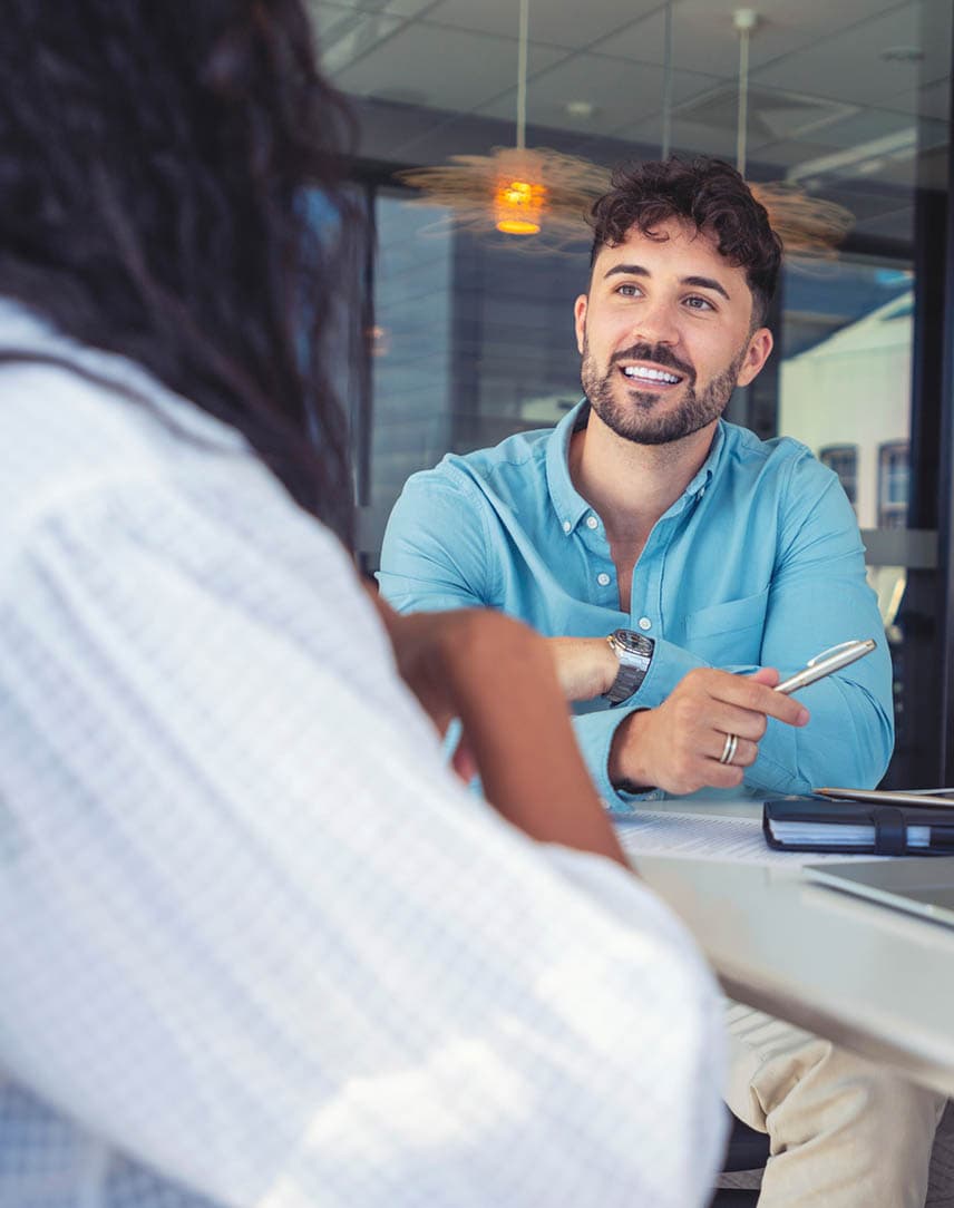 Ein Mann im blauen Hemd spricht an einem Tisch mit einer Kundin, auf dem Tisch steht ein Notebook mit Diagrammen.