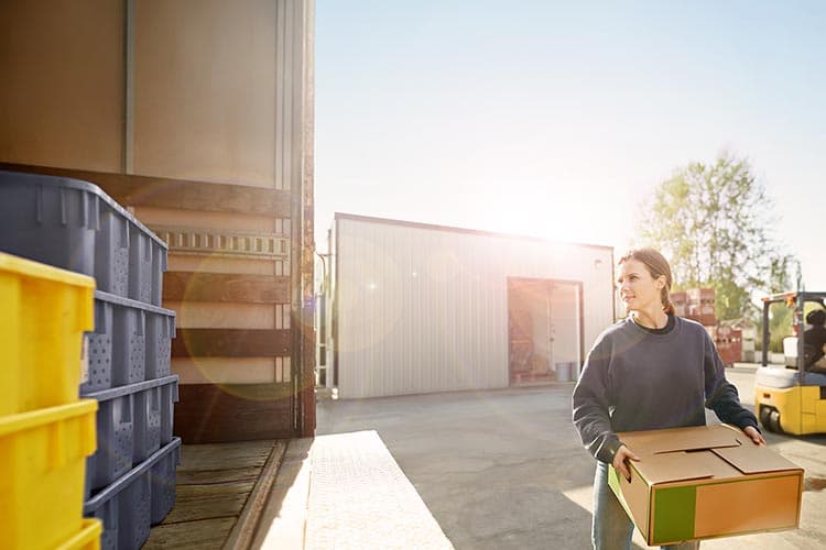 Eine junge Frau räumt einen Karton in einen LKW, in dem Kisten stehen, im Hintergrund ein Gabelstapler, die Sonne scheint.