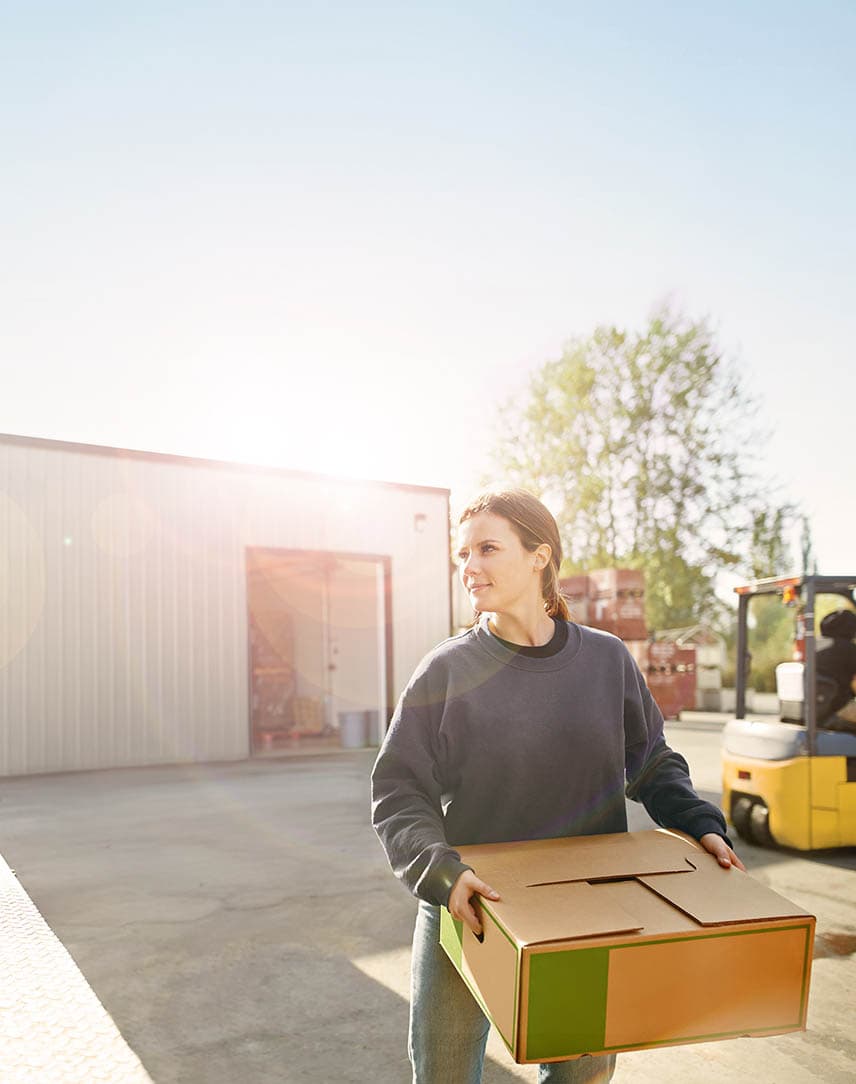 Eine junge Frau räumt einen Karton in einen LKW, in dem Kisten stehen, im Hintergrund ein Gabelstapler, die Sonne scheint.