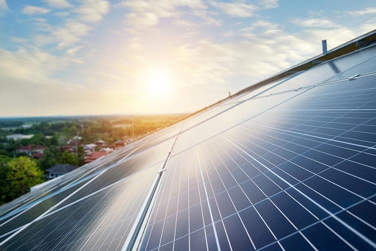 PV-Module auf einem Dach, das von der Sonne bei blauem Himmel beschienen wird, im Hintergrund Landschaft mit Siedlung