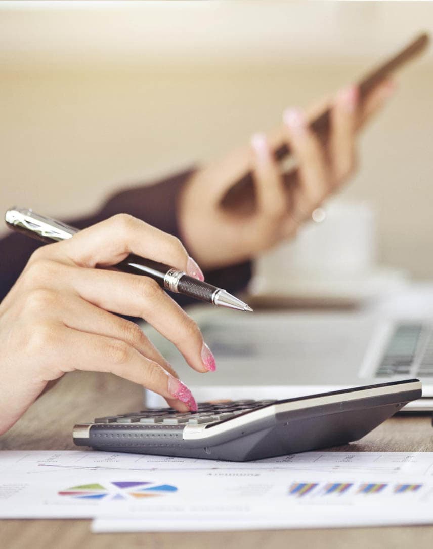 Die Hände einer Person, die ein Handy hält, mit der anderen Hand einen Taschenrechner bedient und einen Stift hält, davor ein Laptop und eine Brille sowie Unterlagen mit Diagrammen