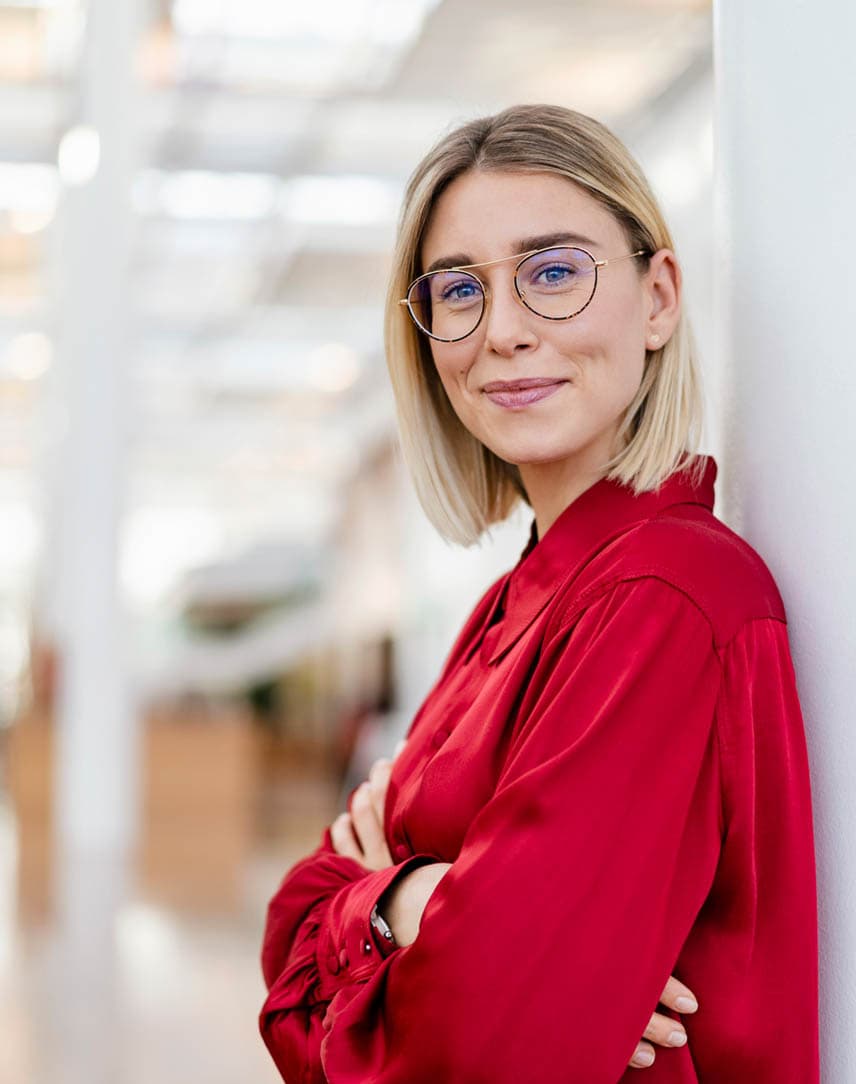 Eine junge Frau mit Brille und in rotem Oberteil lehnt an einer Wand in einem großen, hellen Raum und lächelt.