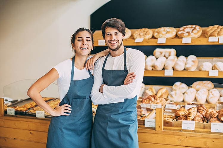 Eine junge Bäckerin und ein junger Bäcker stehen nebeneinander vor einer Brotauslage in einer Bäckerei und lächeln.