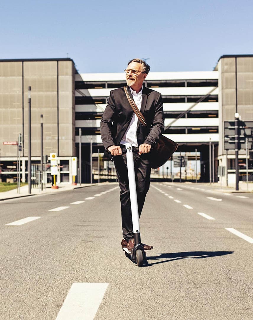 Ein Mann im Anzug fährt mit Tasche auf einem E-Roller auf der Straße aus einem Werksgebäude heraus.