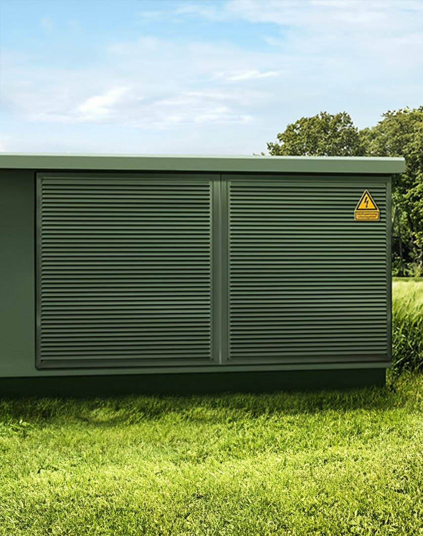 Eine grüne Mittelspannungsanlage auf grüner Wiese, im Hintergrund eine Siedlung unter hellblauem Himmel