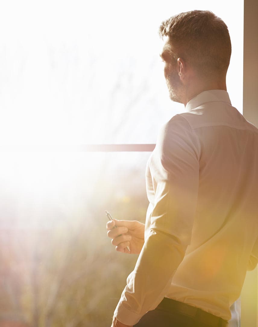 Ein Geschäftsmann guckt aus einem Bürofenster, draußen scheint die Sonne.