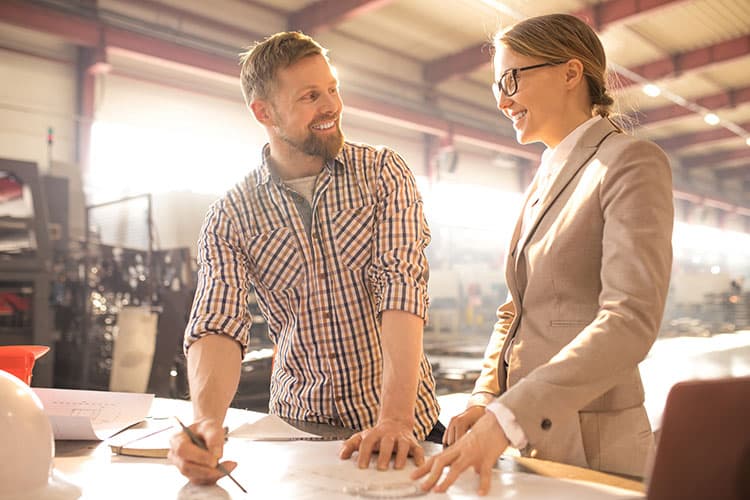 Ein Handwerker im karierten Hemd und eine Frau in Geschäftskleidung stehen in einer Werkstatt, besprechen einen Plan und lächeln.