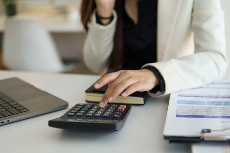 Eine Frau tippt an einem Schreibtisch etwas in einen Taschenrechner, daneben ein Klemmbrett mit Bilanz sowie ein Laptop und außerdem ein Notizbuch