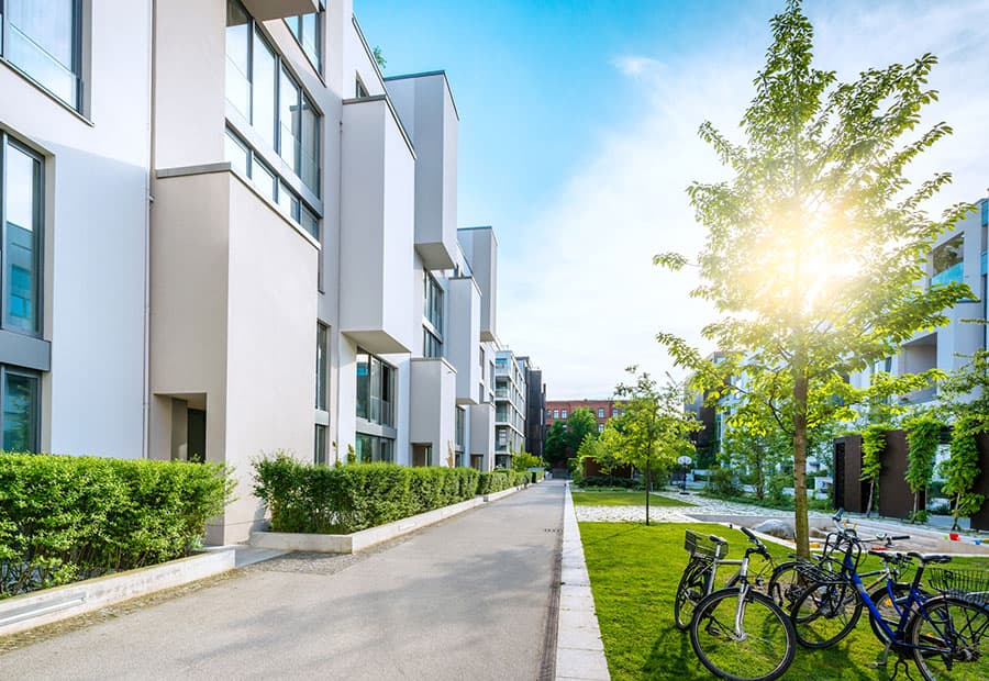 Eine Anlage mit weißen Mehrfamilienhäusern, im Vordergrund ein begrünter Weg und Fahrräder