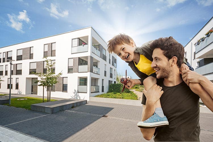 Ein Vater mit einem Jungen auf seinen Schultern, der mit einem Flugzeug spielt, im Hintergrund weiße moderne Wohnblöcke