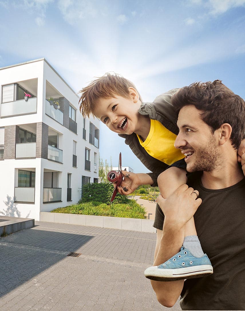 Ein Vater mit einem Jungen auf seinen Schultern, der mit einem Flugzeug spielt, im Hintergrund weiße moderne Wohnblöcke
