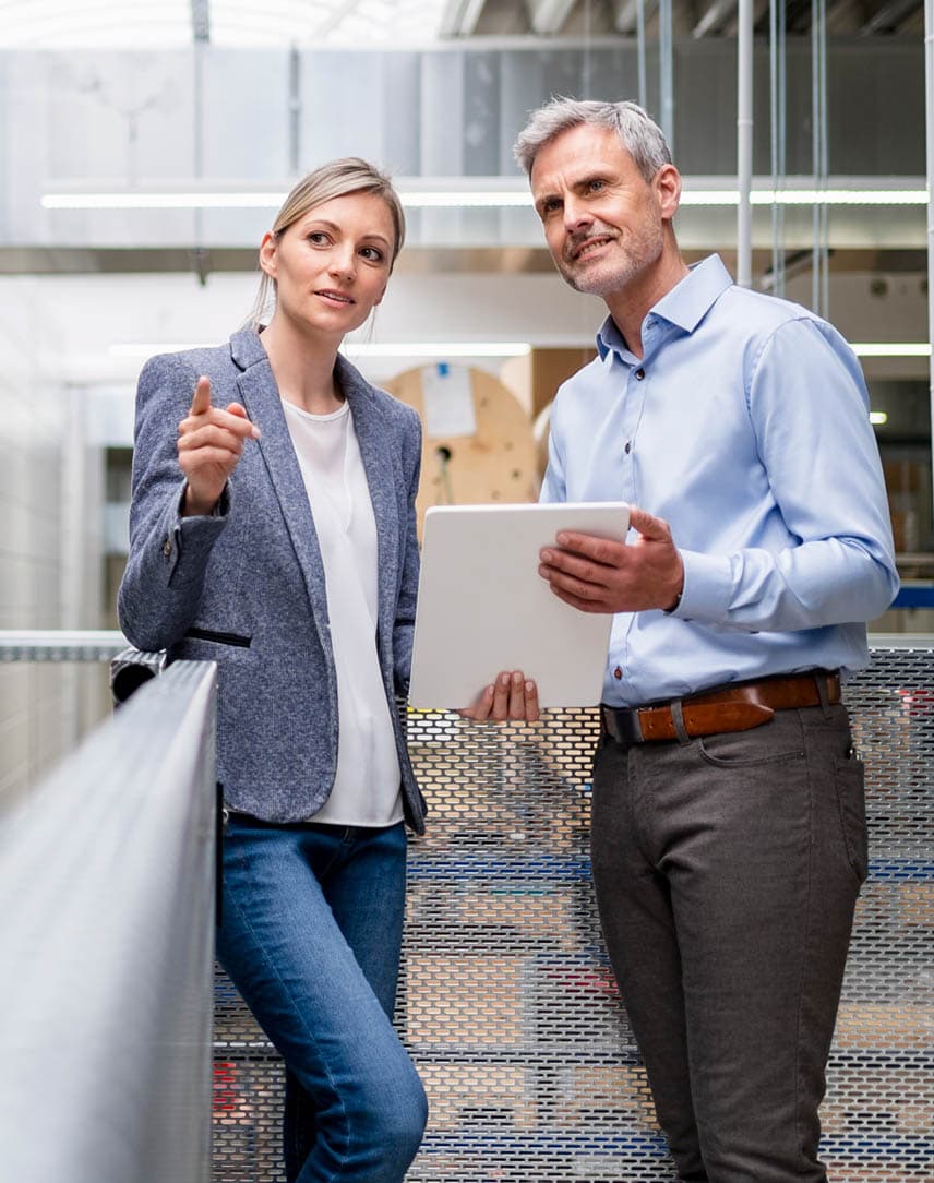 Eine Frau, die auf etwas zeigt, und ein Mann, der ein Tablet hält, besprechen etwas in einer Werkhalle