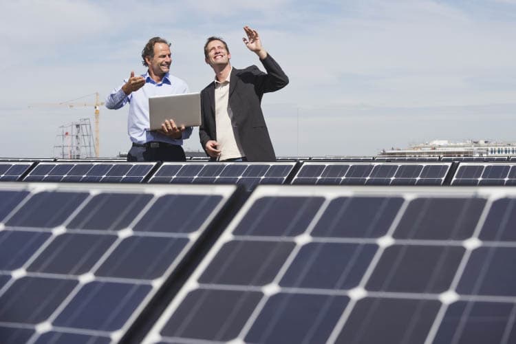 Zwei Männer, von denen einer ein Notebook hält, stehen hinter PV-Modulen und reden miteinander.