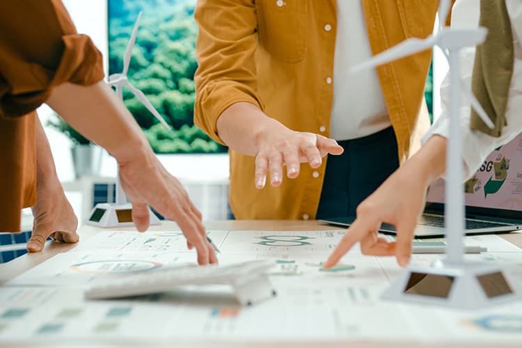 Mehrere Personen zeigen mit den Händen auf Pläne und Modelle von Windkraftanlagen auf einem Tisch