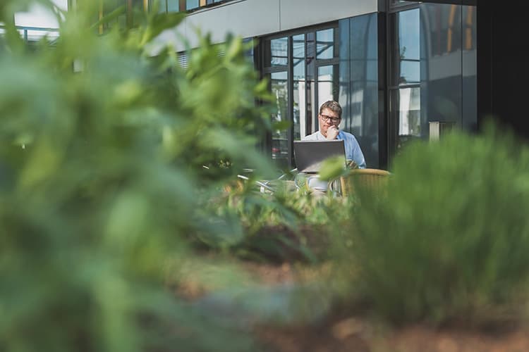 Person sitzt mit Laptop auf einer Bank vor einem modernen Glasgebäude, umgeben von Pflanzen