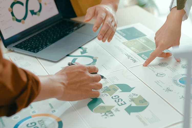 Zwei Personen zeigen mit Händen auf Papiere mit Diagrammen und Symbolen neben einem geöffneten Laptop