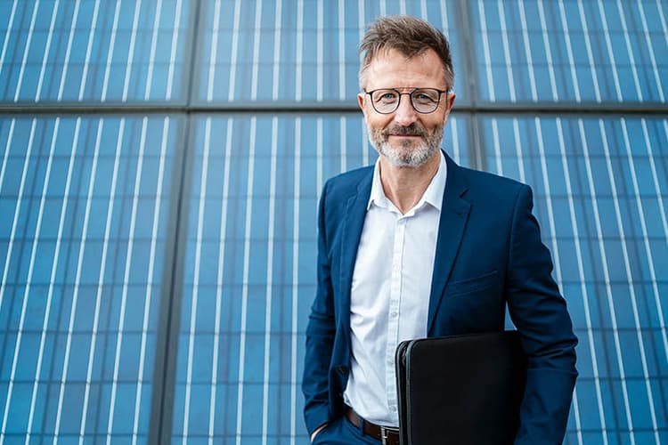Ein Mann in einem dunkelblauen Anzug und weißem Hemd steht vor einer großen Solarzellenwand und hält eine schwarze Mappe unter dem Arm.