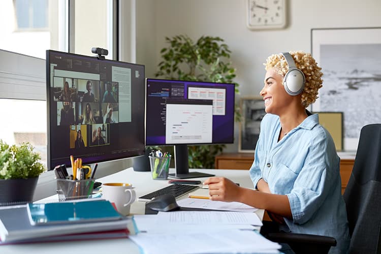 Person mit Kopfhörern sitzt an Schreibtisch mit zwei Monitoren, auf einem ist eine Videokonferenz zu sehen