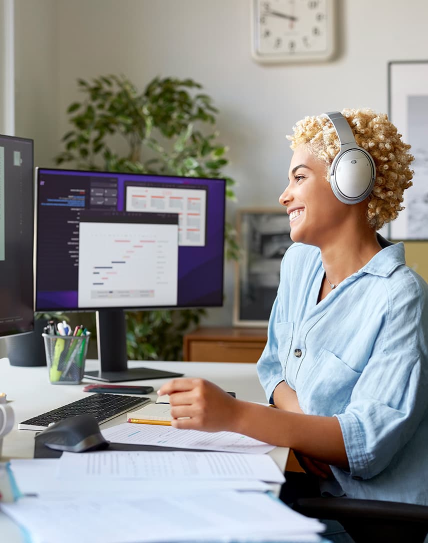 Person mit Kopfhörern sitzt an Schreibtisch mit zwei Monitoren, auf einem ist eine Videokonferenz zu sehen