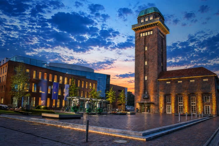Beleuchteter historischer Turm neben modernem Gebäude bei Sonnenuntergang mit bewölktem Himmel