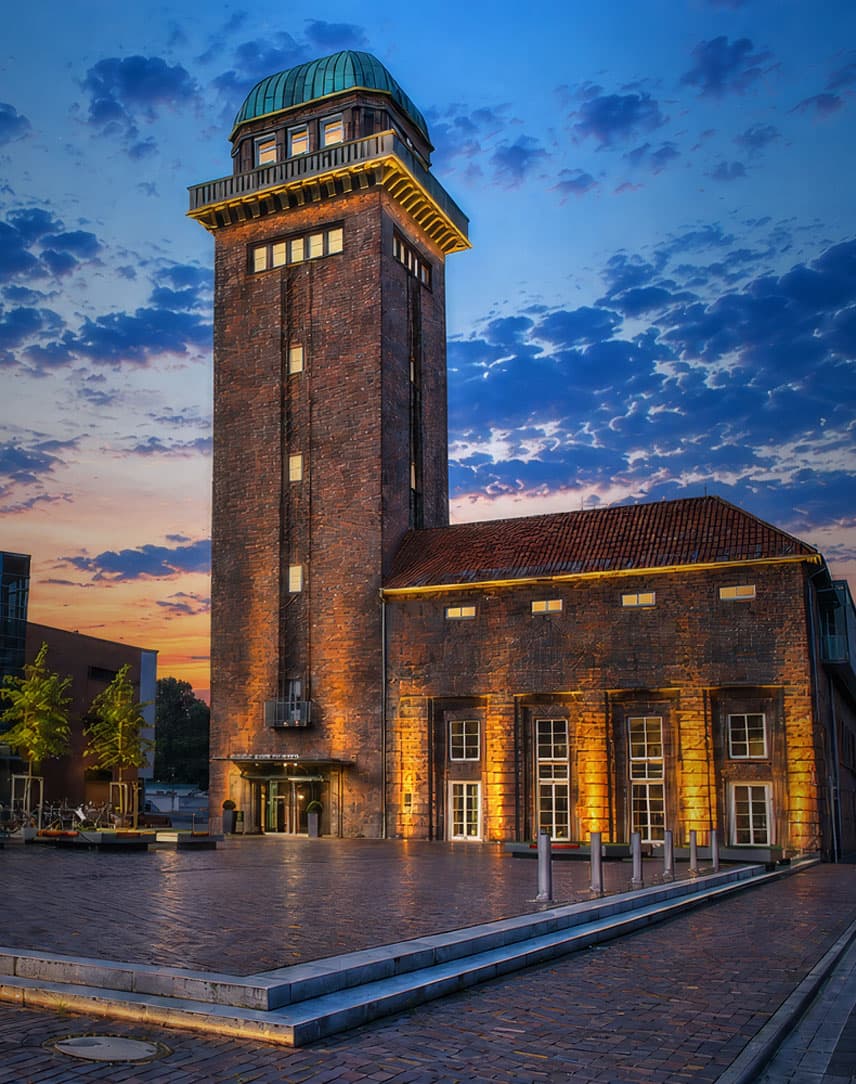 Beleuchteter historischer Turm neben modernem Gebäude bei Sonnenuntergang mit bewölktem Himmel