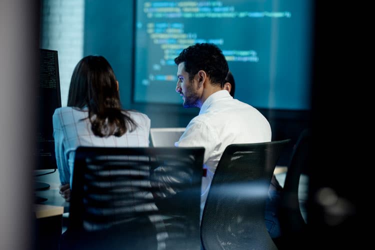 Eine Frau und ein Mann sitzen an einem Arbeitstisch, im Hintergrund ein großer Screen mit Code.