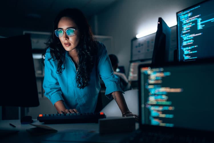 Eine Frau mit Brille arbeitet im Stehen an einem Computer, daneben Bildschirme.