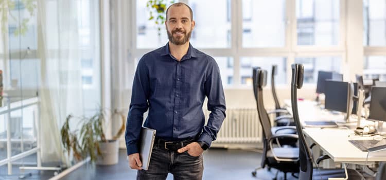 Ein Mann mit Notebook steht in einem Büro, lächelt und hat die linke Hand in der Hosentasche.