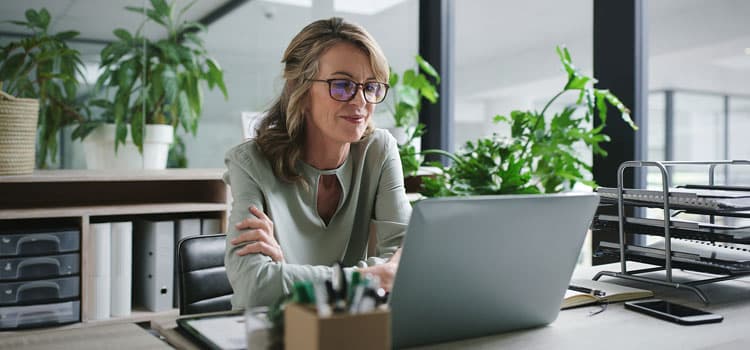 Eine Frau mit Brill stützt sich auf ihren Schreibtisch und sieht sich etwas an einem Notebook an.
