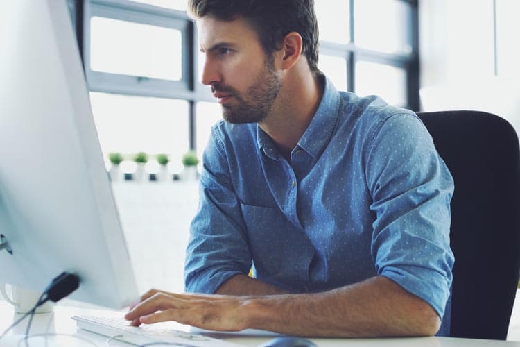 Ein Mann im blauen Hemd guckt konzentriert auf einen Bildschirm am Arbeitstisch.