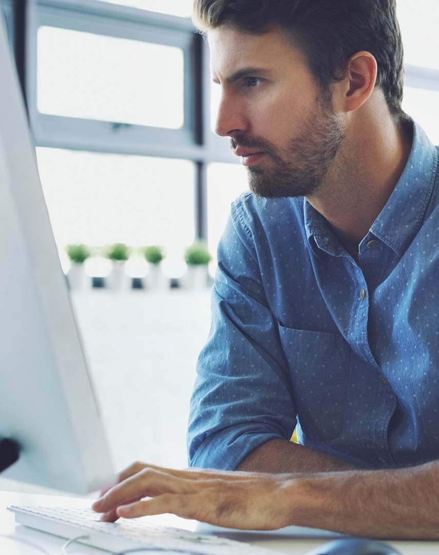 Ein Mann im blauen Hemd guckt konzentriert auf einen Bildschirm am Arbeitstisch.