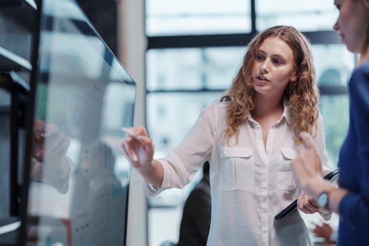 Eine Frau erklärt einer Kollegin etwas an einem großen Bildschirm.