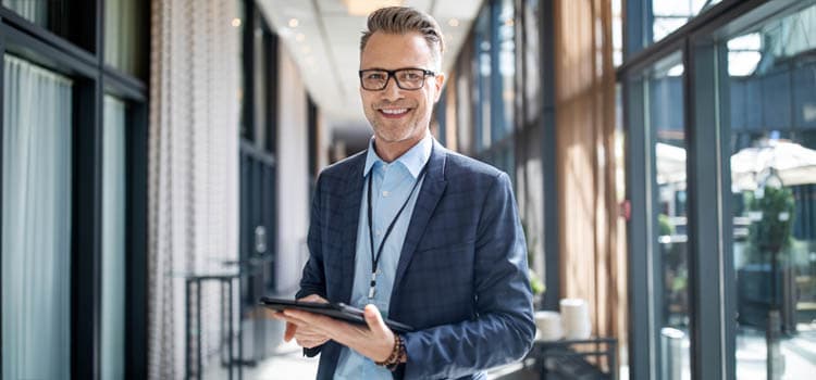 Ein Mann im Jacket mit Tablet und Ausweis, der in einem Gang an einer Fensterfront steht und lächelt