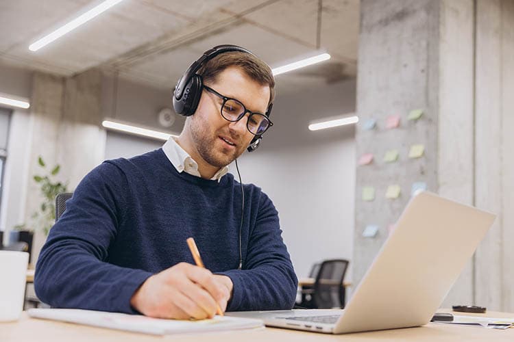 Ein Mann im blauen Pullover mit Headset hört bei einem Meeting zu und macht sich Notizen.