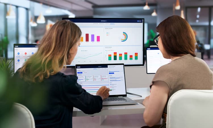 Zwei Frauen, die bei der Arbeit Dashboards auf mehreren Bildschirmen besprechen